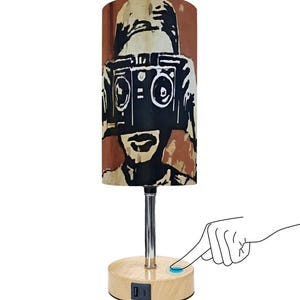 May include: A table lamp with a cylindrical shade featuring a black and brown graphic of a person with a boombox. The lamp has a silver stem and a wooden base with a USB port. The lamp is turned on with a touch sensor.