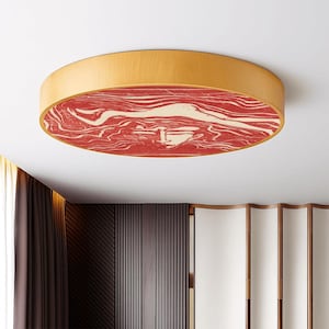 May include: A round flush mount ceiling light fixture. The light has a light wood-colored frame and a red and cream-colored patterned design on the underside. The fixture is mounted on a white ceiling.