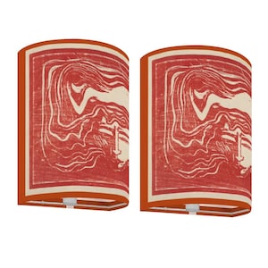 May include: Two cylindrical lampshades with a red and cream-colored design. The design features a stylized image of a person with flowing hair. The lampshades have a rectangular shape and are trimmed with a burnt orange border.