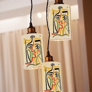 May include: Three hanging pendant lights with a rectangular glass shade. Each shade features a colorful abstract face design in black, red, yellow, and green on a cream background. The fixtures have bronze-colored hardware and black cords.