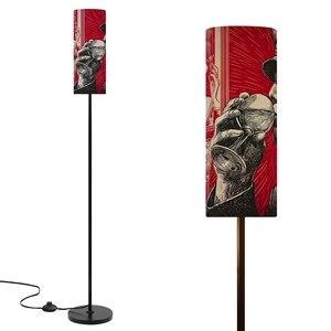 May include: A tall, black floor lamp with a cylindrical lampshade. The lampshade features a red background with a black and white illustration of a person holding a glass. The lamp has a round base.