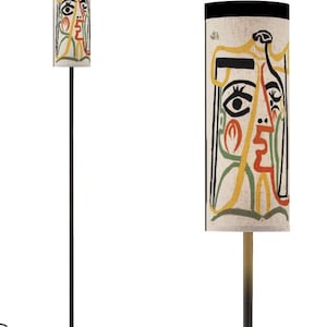 May include: A tall, modern floor lamp with a cylindrical lampshade featuring an abstract face design. The shade has a cream background with black, yellow, red, and green lines. The lamp has a black metal base and pole.