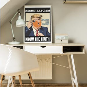 May include: A framed poster with the text "RESIST FASCISM" and "KNOW THE TRUTH" features a caricature of a man with orange skin and blonde hair. The poster also includes the text "ORANGE LIES MATTER". A white desk, chair, and lamp are also visible.