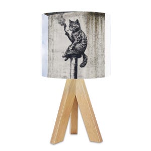 May include: A table lamp with a light wood tripod base and a cylindrical lampshade. The lampshade features a black and white illustration of a cat smoking a pipe. The cat is sitting on a pole, with smoke rising from the pipe.