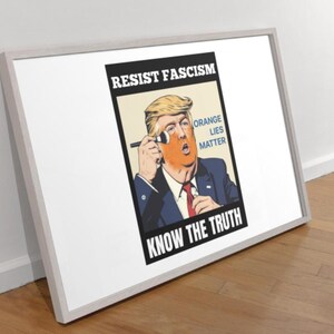 May include: Framed poster with a political cartoon of a man with orange skin applying makeup. Text includes "RESIST FASCISM," "ORANGE LIES MATTER," and "KNOW THE TRUTH." The frame is white, and the poster is leaning against a wall.