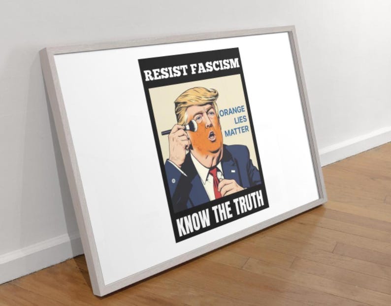 May include: Framed poster with a political cartoon of a man with orange skin applying makeup. Text includes "RESIST FASCISM," "ORANGE LIES MATTER," and "KNOW THE TRUTH." The frame is white, and the poster is leaning against a wall.