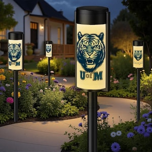 May include: Outdoor pathway lights with a black finish and a glowing panel featuring a tiger head logo and the text "U of M". The lights are set in a garden with colorful flowers and a pathway.