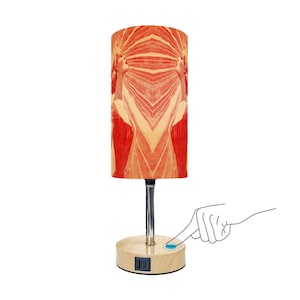 May include: A decorative table lamp with a cylindrical lampshade featuring a symmetrical design in shades of red and beige. The lamp has a chrome stem and a wooden base with a USB port. A finger is touching the power button.