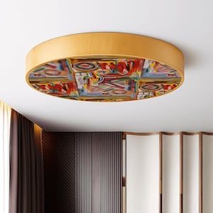 May include: A round flush mount ceiling light fixture with a light wood grain finish. The center of the light features a colorful abstract painting with red, blue, yellow, and green hues. The fixture is mounted on a white ceiling.