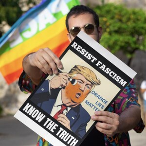 May include: A poster with the text "RESIST FASCISM" and "KNOW THE TRUTH" features a caricature of a man with orange skin. The words "ORANGE LIES MATTER" are also visible. A rainbow flag is in the background.
