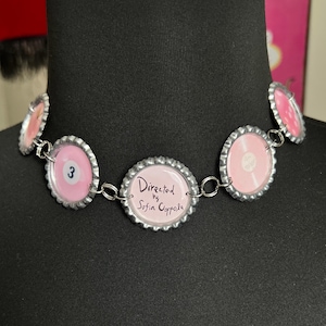 May include: A handmade necklace featuring bottle caps with pink and silver accents. The caps display the number 3, a record design, and the text "Directed by Sofia Coppola". The necklace is displayed on a black mannequin.