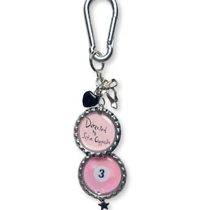 May include: A silver keychain with two pink bottle cap charms. The top charm reads "Directed by Sofia Coppola." The bottom charm features a pink billiard ball with the number 3. A black heart and bow charm are attached.