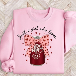 May include: Pink sweatshirt with the text "Just a girl who loves" and a Dr. Pepper can design. The can is decorated with a bow and hearts. The number 23 is on the can.
