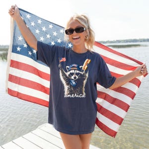 Pu&ograve; includere: Una t-shirt grigio scuro a maniche corte con la grafica di un procione che indossa un berretto e occhiali da sole a forma di cuore, che tiene in mano una piccola bandiera americana e un hot dog, con la scritta "america" in bianco sotto.