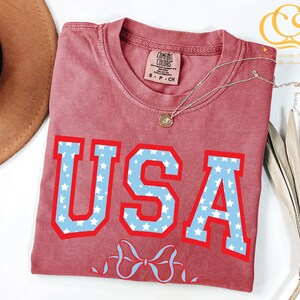 Może przedstawiać: Czerwony T-shirt z biało-niebieskim nadrukiem USA z gwiazdami. Nadruk jest w stylu uniwersyteckim. Koszulka ma postarzony wygląd.