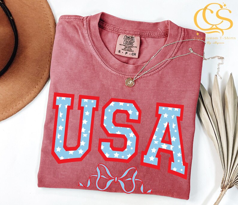 Może przedstawiać: Czerwony T-shirt z biało-niebieskim nadrukiem USA z gwiazdami. Nadruk jest w stylu uniwersyteckim. Koszulka ma postarzony wygląd.