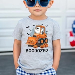 Peut inclure: T-shirt gris clair avec un motif d'Halloween. Il pr&eacute;sente un bulldozer orange avec un fant&ocirc;me, des citrouilles et des chauves-souris. Le texte "BOODOZER" est imprim&eacute; en dessous. Le t-shirt est port&eacute; par un enfant.