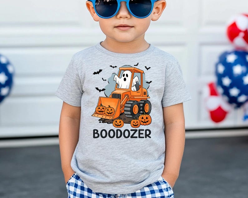 Peut inclure: T-shirt gris clair avec un motif d'Halloween. Il pr&eacute;sente un bulldozer orange avec un fant&ocirc;me, des citrouilles et des chauves-souris. Le texte "BOODOZER" est imprim&eacute; en dessous. Le t-shirt est port&eacute; par un enfant.