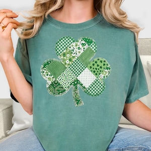 May include: A teal green t-shirt featuring a patchwork shamrock design. The shamrock is composed of various green patterned fabrics, including gingham, floral, and paisley prints. The shirt is a comfortable, casual style.