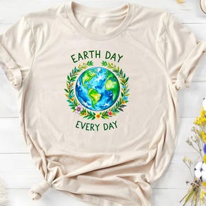 Puede incluir: Una camiseta beige con un gr&aacute;fico verde y azul de la Tierra rodeada de flores y hojas. El texto "Earth Day Every Day" est&aacute; impreso en verde debajo del gr&aacute;fico.