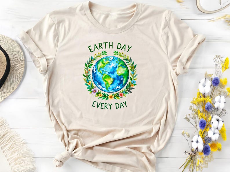 Puede incluir: Una camiseta beige con un gr&aacute;fico verde y azul de la Tierra rodeada de flores y hojas. El texto "Earth Day Every Day" est&aacute; impreso en verde debajo del gr&aacute;fico.