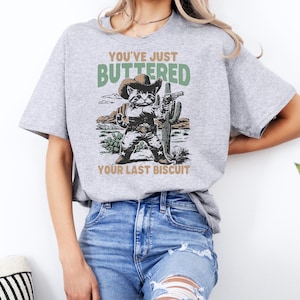 May include: A light gray t-shirt featuring a cartoon cat dressed as a cowboy, holding a gun and standing in front of a cactus. The text on the shirt reads "YOU'VE JUST BUTTERED YOUR LAST BISCUIT."