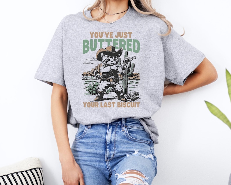 May include: A light gray t-shirt featuring a cartoon cat dressed as a cowboy, holding a gun and standing in front of a cactus. The text on the shirt reads "YOU'VE JUST BUTTERED YOUR LAST BISCUIT."