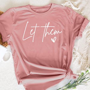 May include: A pink t-shirt with the text "Let Them" in white lettering and a small white heart.