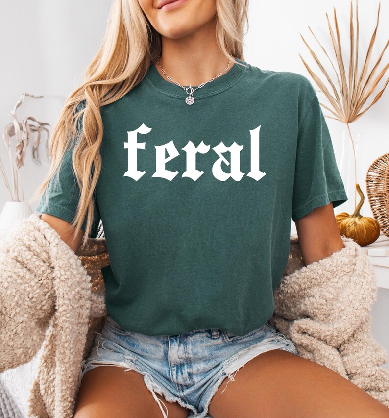 Pode incluir: Uma camiseta verde-azulada com a palavra "feral" impressa em branco em uma fonte g&oacute;tica.