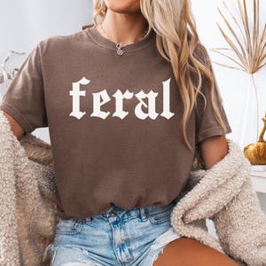 Pode incluir: Uma camiseta marrom com a palavra "feral" impressa em branco em uma fonte g&oacute;tica.