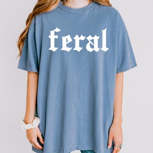 Pode incluir: Uma camiseta azul oversized com a palavra "feral" impressa em branco em uma fonte g&oacute;tica.
