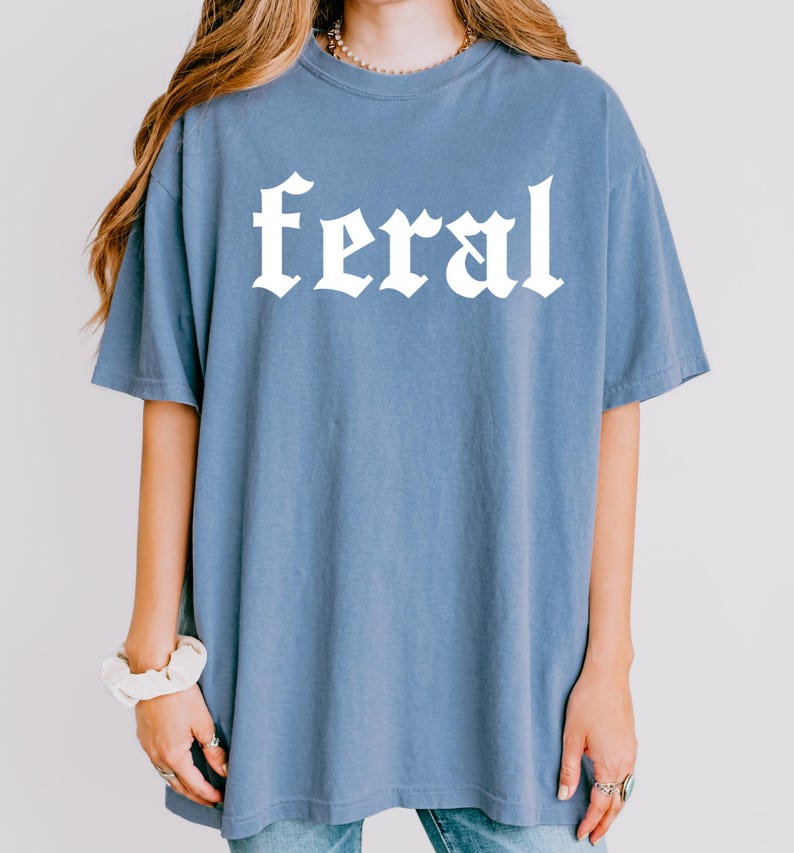Pode incluir: Uma camiseta azul oversized com a palavra "feral" impressa em branco em uma fonte g&oacute;tica.