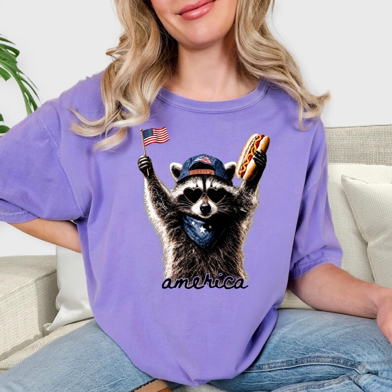 Pu&ograve; includere: Una t-shirt viola chiaro con la grafica di un procione che indossa occhiali da sole a forma di cuore, un berretto da baseball blu e una bandana blu. Il procione tiene una piccola bandiera americana e un hot dog, con la scritta america sotto.