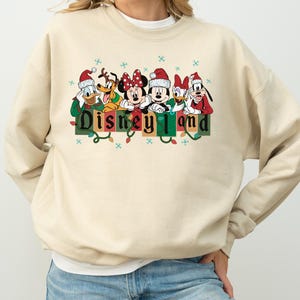 K&ouml;nnte beinhalten: Ein beiges Sweatshirt mit den Disney-Figuren Donald Duck, Pluto, Minnie Maus, Mickey Maus, Daisy Duck und Goofy, alle mit Weihnachtsm&uuml;tzen. Das Wort "Disneyland" ist in gr&uuml;nen und schwarzen Blockbuchstaben mit Lichterketten gedruckt.