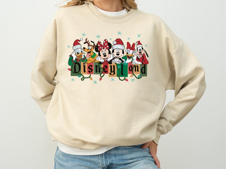 K&ouml;nnte beinhalten: Ein beiges Sweatshirt mit den Disney-Figuren Donald Duck, Pluto, Minnie Maus, Mickey Maus, Daisy Duck und Goofy, alle mit Weihnachtsm&uuml;tzen. Das Wort "Disneyland" ist in gr&uuml;nen und schwarzen Blockbuchstaben mit Lichterketten gedruckt.