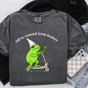 May include: Dark gray t-shirt with a green frog wearing a wizard hat and riding a scooter, holding a four-leaf clover. Text reads "Off to commit Irish foolery". The shirt is paired with blue jeans and a phone with a checkered case.