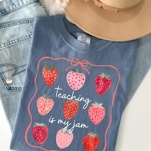 May include: A blue Comfort Colors t-shirt with a strawberry design and the text "Teaching is my jam." The shirt is paired with blue jeans and a tan hat. The strawberries are red and pink.