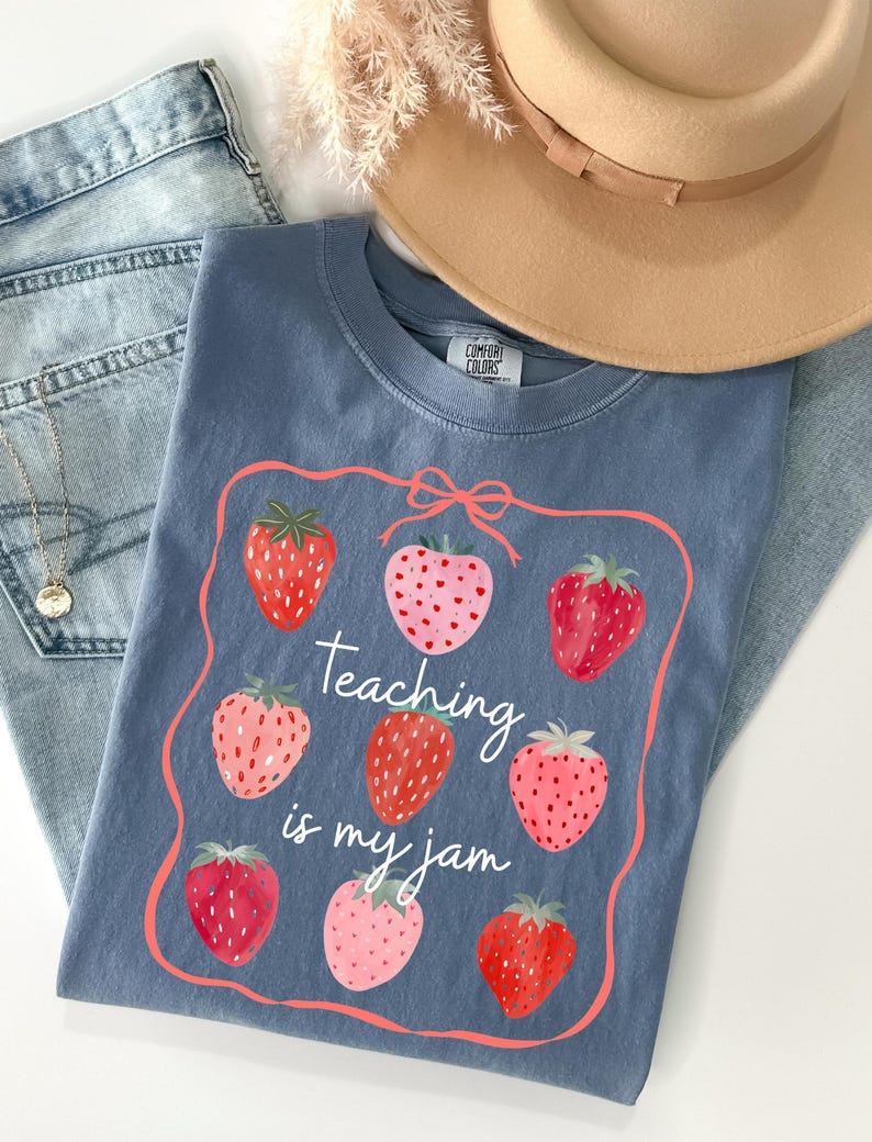 May include: A blue Comfort Colors t-shirt with a strawberry design and the text "Teaching is my jam." The shirt is paired with blue jeans and a tan hat. The strawberries are red and pink.