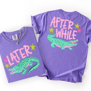May include: Two purple t-shirts with a graphic design featuring a green alligator and the words "LATER" and "AFTER WHILE" in pink. The shirts have a crew neck and short sleeves. One shirt has a silver chain necklace.