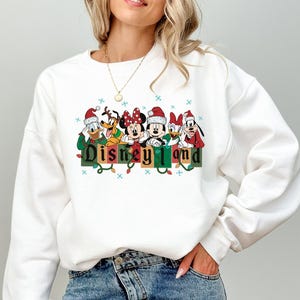 K&ouml;nnte beinhalten: Wei&szlig;es Sweatshirt mit einem festlichen Aufdruck von Disney-Figuren mit Weihnachtsm&uuml;tzen &uuml;ber dem Wort "Disneyland". Das Design umfasst Micky und Minnie Maus, Donald Duck und Goofy. Das Sweatshirt hat ein Feiertags-Thema.
