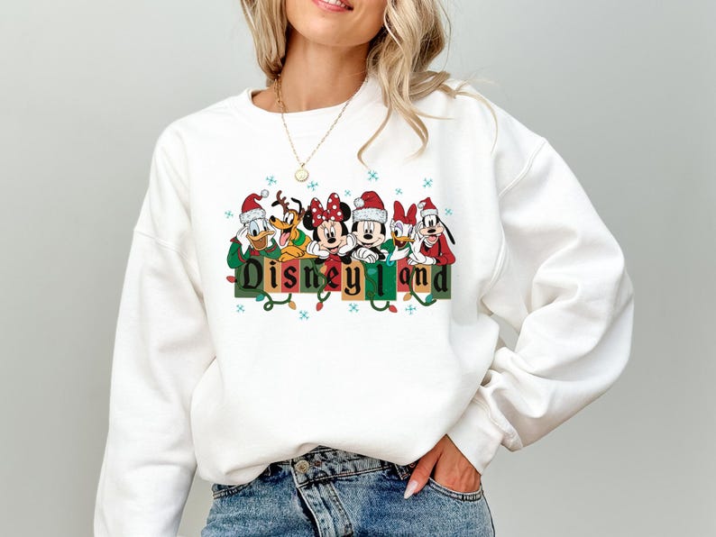 K&ouml;nnte beinhalten: Wei&szlig;es Sweatshirt mit einem festlichen Aufdruck von Disney-Figuren mit Weihnachtsm&uuml;tzen &uuml;ber dem Wort "Disneyland". Das Design umfasst Micky und Minnie Maus, Donald Duck und Goofy. Das Sweatshirt hat ein Feiertags-Thema.