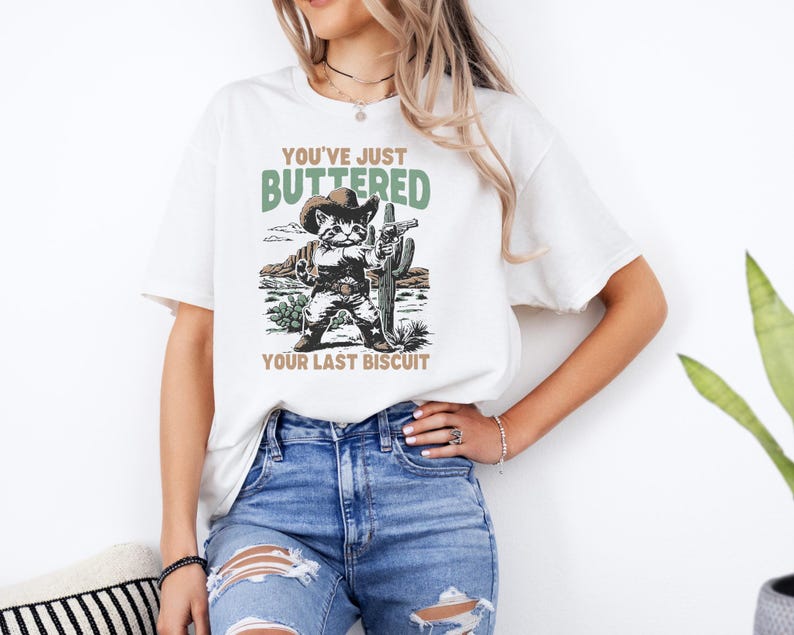 May include: White t-shirt featuring a cartoon raccoon dressed as a cowboy, holding a gun, and standing in front of a desert scene. The text on the shirt reads "YOU'VE JUST BUTTERED YOUR LAST BISCUIT."