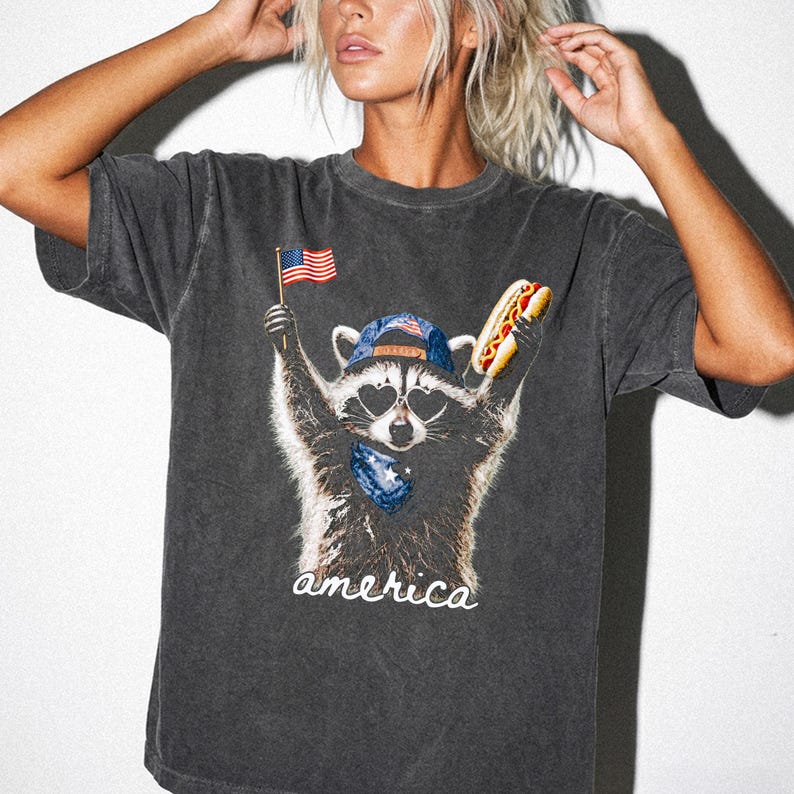 Pu&ograve; includere: Una t-shirt grigio antracite a maniche corte con la grafica di un procione che indossa occhiali da sole a forma di cuore, un berretto da baseball e una bandana. Il procione tiene in mano una bandiera americana e un hot dog. La scritta america &egrave; bianca.