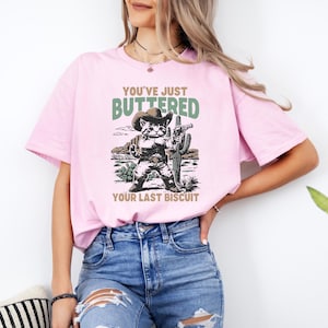 May include: A light pink t-shirt featuring a graphic of a cat in a cowboy hat and holding a gun, with the text "YOU'VE JUST BUTTERED YOUR LAST BISCUIT" in green and brown. The cat is standing in front of a desert scene.