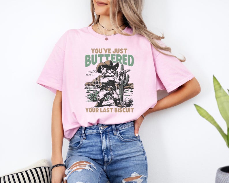 May include: A light pink t-shirt featuring a graphic of a cat in a cowboy hat and holding a gun, with the text "YOU'VE JUST BUTTERED YOUR LAST BISCUIT" in green and brown. The cat is standing in front of a desert scene.