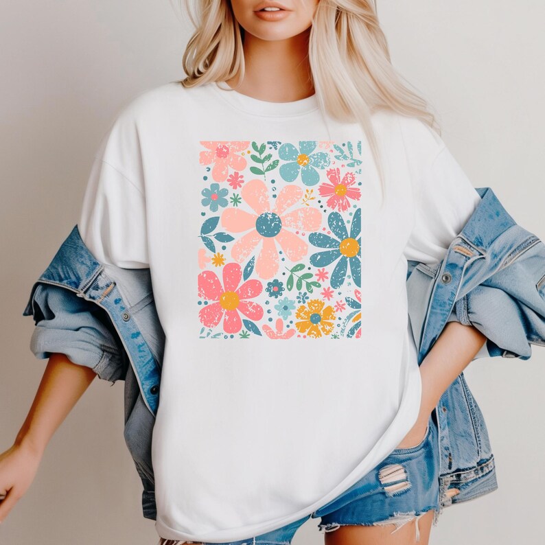 以下が含まれることがあります： カラフルな花柄がプリントされた白いTシャツ。ピンク、青、黄色の大きな花と緑の葉やアクセントが描かれています。ゆったりとしたフィット感で、カジュアルな着こなしに最適です。