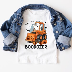 Peut inclure: T-shirt blanc avec un motif sur le th&egrave;me d'Halloween. Le dessin repr&eacute;sente un bulldozer orange avec un fant&ocirc;me, des citrouilles et des chauves-souris. Le mot "BOODOZER" est imprim&eacute; en dessous. Une veste en jean bleue est drap&eacute;e sur le t-shirt.