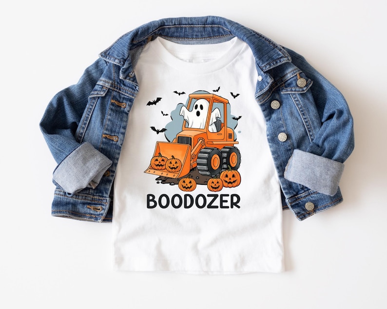 Peut inclure: T-shirt blanc avec un motif sur le th&egrave;me d'Halloween. Le dessin repr&eacute;sente un bulldozer orange avec un fant&ocirc;me, des citrouilles et des chauves-souris. Le mot "BOODOZER" est imprim&eacute; en dessous. Une veste en jean bleue est drap&eacute;e sur le t-shirt.