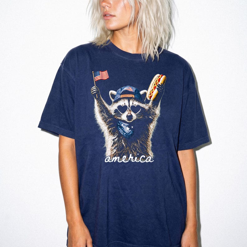 Pu&ograve; includere: Una t-shirt blu navy con la grafica di un procione che indossa un berretto da baseball e occhiali da sole a forma di cuore. Il procione tiene in mano una piccola bandiera americana e un hot dog, con la scritta america in corsivo bianco.