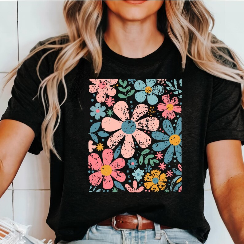 以下が含まれることがあります： カラフルな花柄が特徴の黒いTシャツ。ピンク、青、黄色の大きな花が、ヴィンテージ風の風合いで描かれています。緑の葉と小さな花がパターンを完成させています。